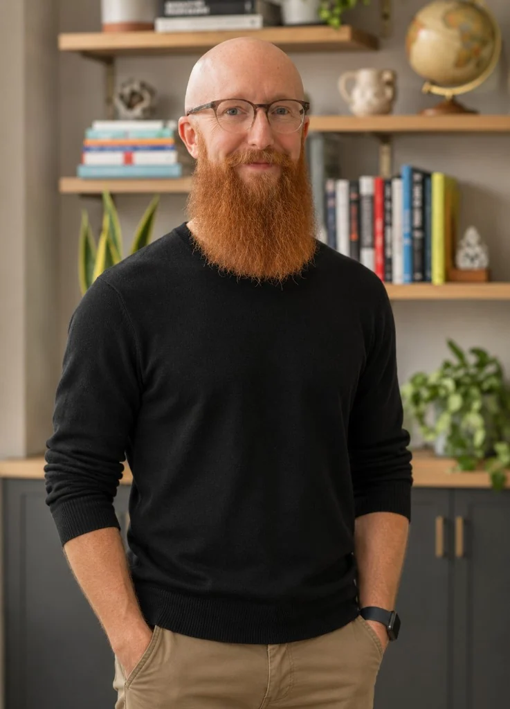 Cameron Sanderson, bald with a red beard and glasses, wearing a black sweater and khaki pants, standing with his hands in his pockets in front of bookshelves.